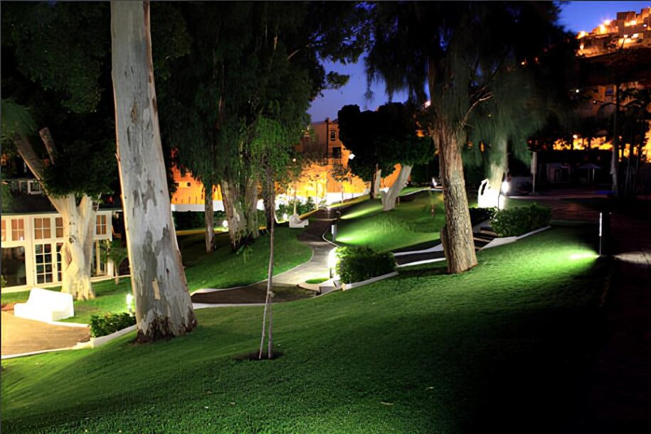 Foto de jardines del club de noche. Varios árboles y césped. Un sendero curvilíneo recorre el jardín. Luces que iluminan el escenario.