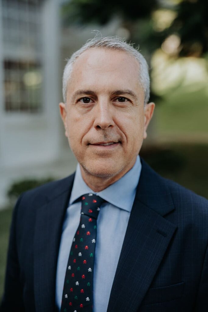 Foto de medio cuerpo superior de Alejandro Tosco. Pelo canoso, ojos negros. Lleva chaqueta y corbata azul marino. Camisa azul claro. Al fondo, borroso, se adivina jardín y parte de la casa.