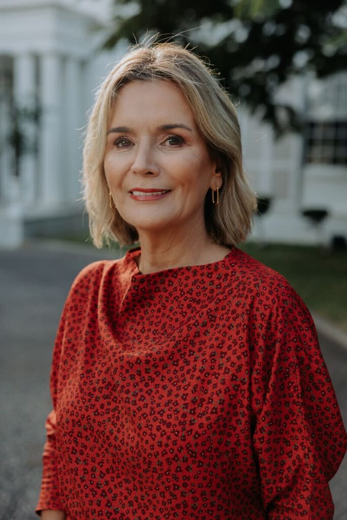 Foto de medio cuerpo superior de María Cristina Sánchez Mayor. Señora de mediana edad. Pelo rubio, ojos negros. Lleva elegante traje rojo. Al fondo y, algo borrosa, se ve parte de la fachada del club con vegetación ante ella, en la parte superior derecha.