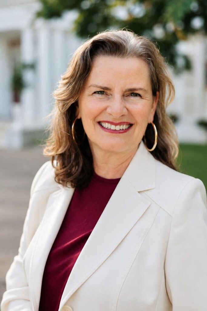 Foto de medio cuerpo superior de Dolores Rodríguez de Azero. Pelo castaño, ojos azules, amplia sonrisa. Lleva grandes pendientes tipo aro. Elegante chaqueta blanca sobre blusa rojo granate. Al fondo, algo borroso, se observa parte de la fachada del club y cierta vegetación.