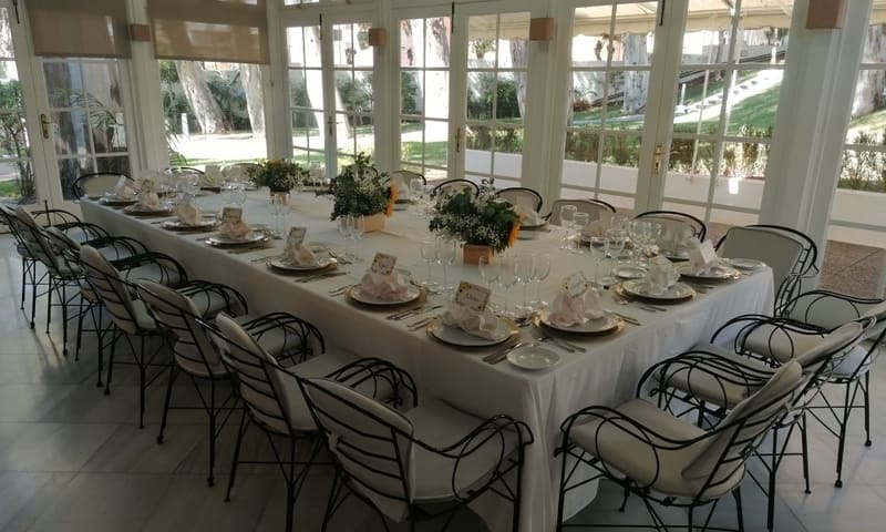 Mesa con mantel blanco para 20 comensales. En zona acristalada con maderas blancas. Se ven jardines a través de las ventanas.
