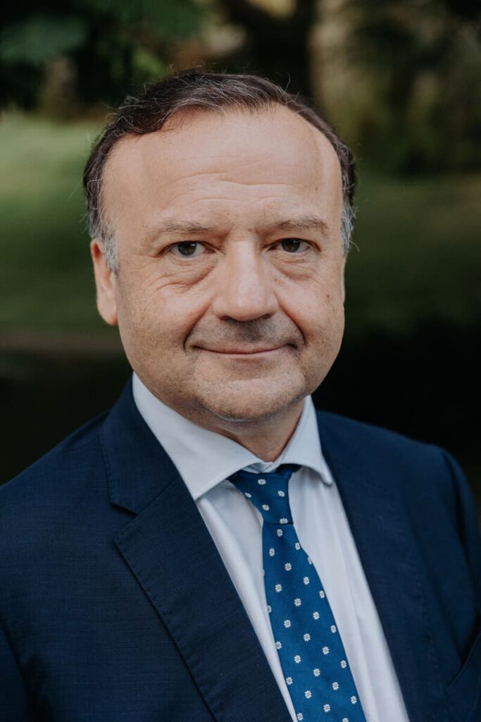 Foto de medio cuerpo de Jesús Barragán. Pelo negro, sonrisa. Chaqueta azul marino. Camisa blanca. Corbata Azul más claro con moteado blanco. Al fondo, muy borroso, jardines.