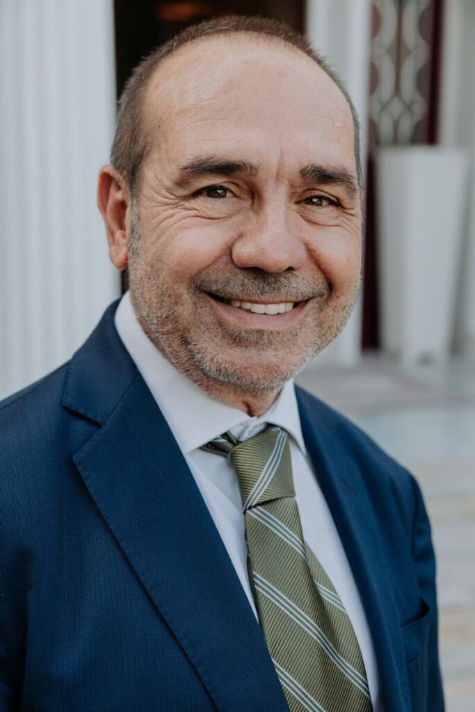 Foto de José Luis Batista, de medio cuerpo superior. Poco pelo, amplia sonrisa. Barba corta. Chaqueta azul marino y corbata verde oliva. Camisa blanca. Al fondo parte de fachada del club, blanca.