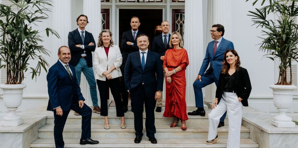 Miembros de la junta posando en las escaleras de la fachada del club. Son 9. Visten elegantemente. Se disponen en tres filas. A ambos lados del conjunto, dos macetas con plantas verdes. Tras el grupo, la puerta principal de entrada al club. Fachada de color blanco.
