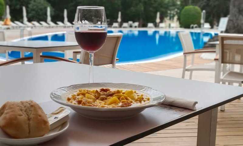Mesa en primer término. Sobre ella un plato con comida y otro pequeño con un pan y una pieza de mantequilla. Copa de vino tinto. Al fondo destaca el azul de la piscina y vegetación.