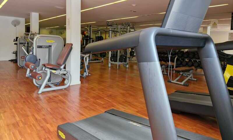 Gimnasio. En primer término máquina de running, gris oscuro. En segundo plano otros aparatos tradicionales de gimnasios. Suelo de parquet.