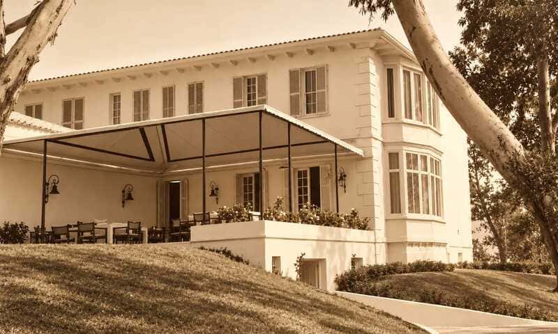 Foto de fachada club oliver parcial. Tomada desde un ángulo. En primer plano una terraza cuberta, sustentada con pilares metálicos de color negro. La foto es de color sepia. El edificio es blanco. Abundan las ventanas. Mansión de estilo rural británico.