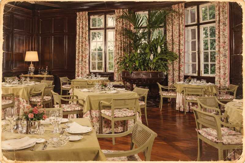Foto antigua de comedor noble. Mesas cuadradas de color verde oliva claro, ya vestidas y con platos. Al fondo ventanas blancas de madera. Las paredes del comedor son de madera, en color caoba.