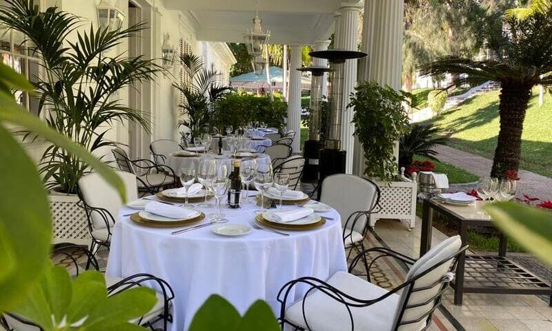Mesas redondas bajo porche de mansión club oliver. Manteles blancos, sillas de metal negras, vestidas de blanco. Cuatro platos en cada mesas, junto a copas de vino vacías. Abundante vegetación.