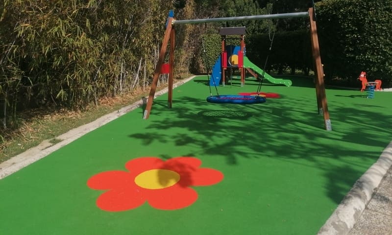 Parque infantil. Suelo verde acolchado, con flor roja y amarilla. Un columpio, un tobogán y un caballito con resorte. Alrededor, vegetación verde.